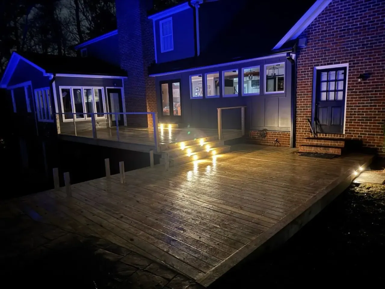 Deck at night lit by recessed riser lights with blue accent uplighting on the house